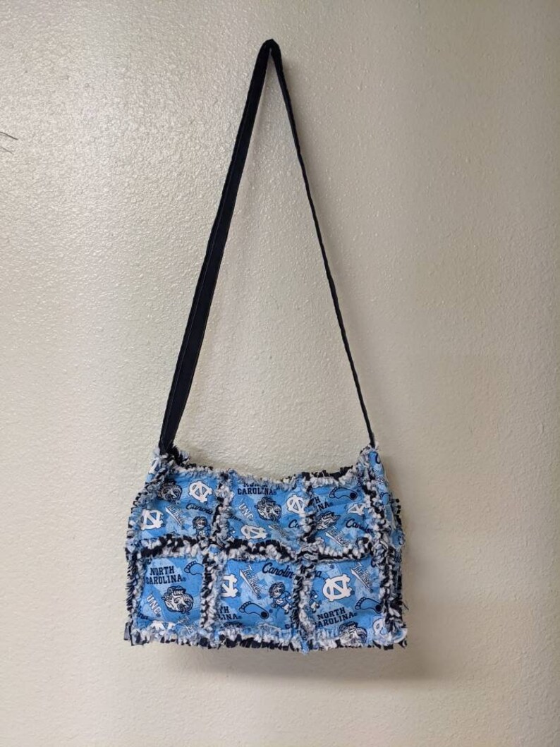 May include: A blue and white patchwork tote bag with a black strap. The bag features a repeated pattern of the University of North Carolina Tar Heels logo and the words "North Carolina".