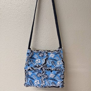 May include: A blue and white fabric crossbody bag with a frayed edge and a black strap. The bag features a repeated pattern of the UNC Tar Heels logo and the words "North Carolina" and "UNC".