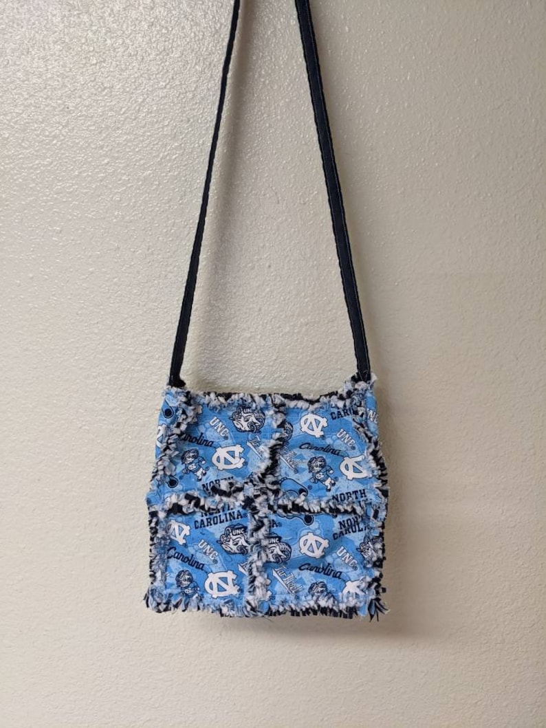 May include: A blue and white fabric crossbody bag with a frayed edge and a black strap. The bag features a repeated pattern of the UNC Tar Heels logo and the words "North Carolina" and "UNC".