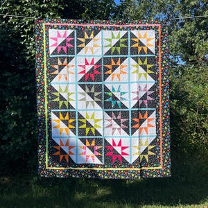May include: A colorful handmade quilt with a star pattern. The quilt features a black background with colorful stars, and a central design of star blocks in various colors like pink, orange, and green. The quilt is hanging outside.