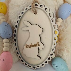 Peut inclure: Ornement de Pâques en bois avec un lapin et le nom "Raphael" gravé. L'ornement ovale a un bord festonné et une ficelle de jute. Des œufs de Pâques aux couleurs pastel sont visibles en arrière-plan.
