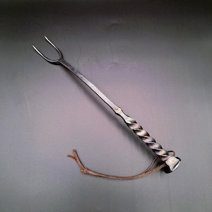 May include: Hand-forged metal grilling fork with a twisted handle and two tines. The fork has a dark, metallic finish and a leather loop for hanging. The design is rustic and handcrafted, suitable for outdoor cooking.
