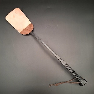 May include: Hand-forged metal spatula with a copper-colored blade and a twisted handle. The spatula has a dark finish and a leather loop at the end of the handle. The blade is rectangular, and the handle is long and slender.