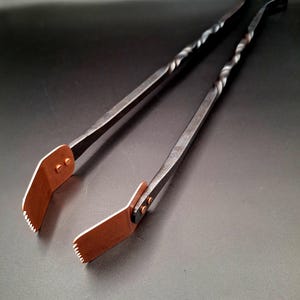 Puede incluir: Pinzas de metal forjadas a mano con un diseño de mango retorcido. Las pinzas tienen puntas de color cobre con un borde dentado. El metal es oscuro y el diseño general es rústico y funcional.