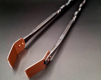 May include: Hand-forged metal tongs with a twisted handle design. The tongs feature copper-colored tips with a serrated edge. The metal is dark, and the overall design is rustic and functional.