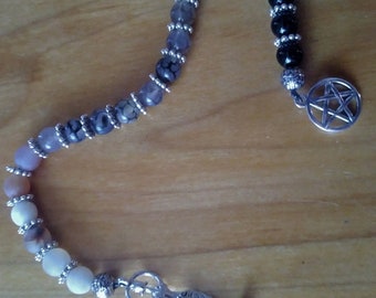 May include: A beaded bracelet featuring a gradient of smooth round beads transitioning from white and tan to grey, black, and dark patterned stones. The piece is accented with silver-toned metal spacers, a silver pentagram charm, and a metal toggle clasp.