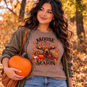 May include: A taupe t-shirt featuring a graphic of a moose wearing a plaid scarf, surrounded by sunflowers, pumpkins, and autumn leaves. The text reads "MOOSE Be Cozy SEASON" in a dark brown font. The shirt is worn under a dark olive jacket.