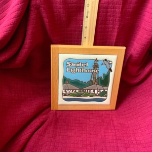 May include: Square ceramic tile with a wooden frame. The tile features a colorful illustration of the Sanibel Lighthouse with the text "Sanibel Lighthouse". A ruler is placed behind the tile.