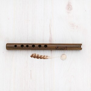May include: A brown wooden flute with six finger holes and a mouthpiece. The flute is labeled "Lupaca" and is lying on a white wooden surface.