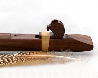 May include: A dark brown wooden flute featuring a carved bird-shaped block secured with a light tan leather strap. A single long feather with brown and white mottled patterns lies horizontally beneath the body of the musical instrument.
