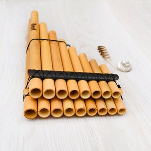 May include: A wooden panpipe with 14 pipes bound together with black string. The panpipe is made of natural bamboo and is lying on a light wood surface.