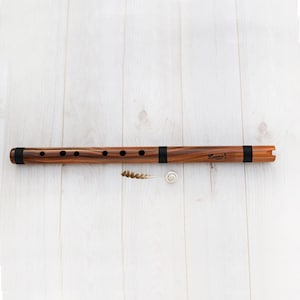 May include: A wooden flute with a dark brown finish and black bands. The flute has several finger holes and the text "Lumen" is visible. A cleaning brush and a small white spiral are next to the flute.