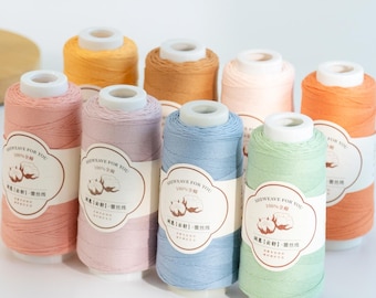 May include: A collection of colorful spools of thread in various pastel shades, including pink, blue, and green. Each spool has a white label with text and a cotton plant illustration. The spools are arranged on a white surface.