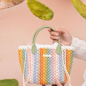 May include: A handmade, colourful crochet bag with a light green handle. The bag features a mix of pastel colours, including orange, yellow, pink, and blue. The word "handmade" is on a small tag on the bag. The background includes green cactus leaves and a pink wall.