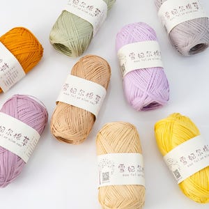 以下が含まれることがあります： 黄色、オレンジ、緑、ベージュ、ラベンダー、グレーの色合いのカラフルな糸玉の詰め合わせ。各糸玉には中国語の文字が書かれた白いラベルが付いています。編み物や編み物プロジェクトに最適です。