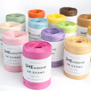 Op de afbeelding: Een verzameling meerkleurige raffia garenspoelen met het label SHEweave. Elke spoel heeft een witte papieren band met de tekst SHEweave, 150g / 300m en Chinese karakters. De garenkleuren zijn onder andere roze, paars, geel, groen, blauw, oranje en bruin.