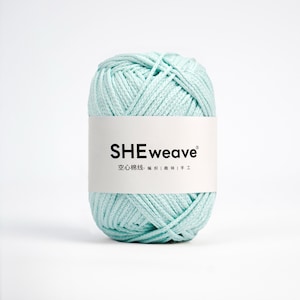 May include: A ball of light mint green yarn wrapped with a white paper label featuring the black text SHEweave and Chinese characters. The yarn has a smooth, cord-like texture and is wound into a compact, cylindrical shape for crafting.