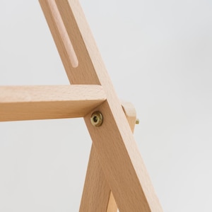 May include: A light-colored wooden easel frame featuring a horizontal support bar and a vertical frame member connected by a circular brass-toned metal bolt. The structure is composed of smooth, unfinished wood with visible grain and clean joints.