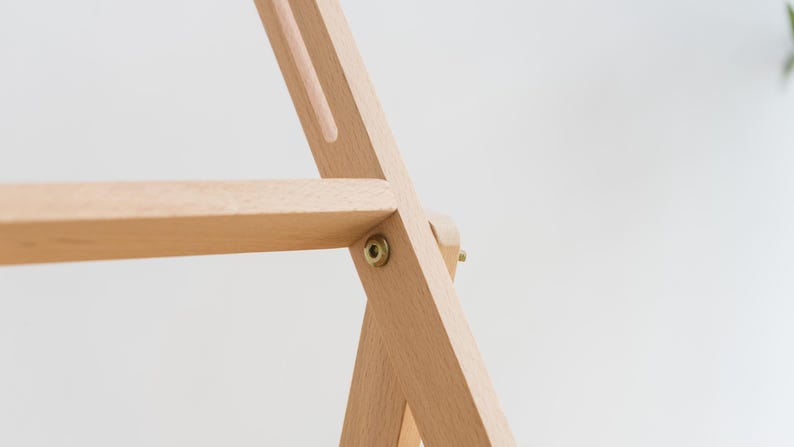 May include: A light-colored wooden easel frame featuring a horizontal support bar and a vertical frame member connected by a circular brass-toned metal bolt. The structure is composed of smooth, unfinished wood with visible grain and clean joints.