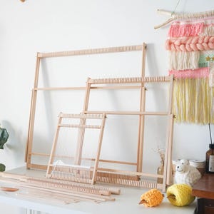 May include: Three light-colored wooden weaving looms of varying sizes are arranged on a white surface. The looms feature vertical notches along the top and bottom bars for threading. Several wooden weaving tools and balls of yarn are placed nearby.