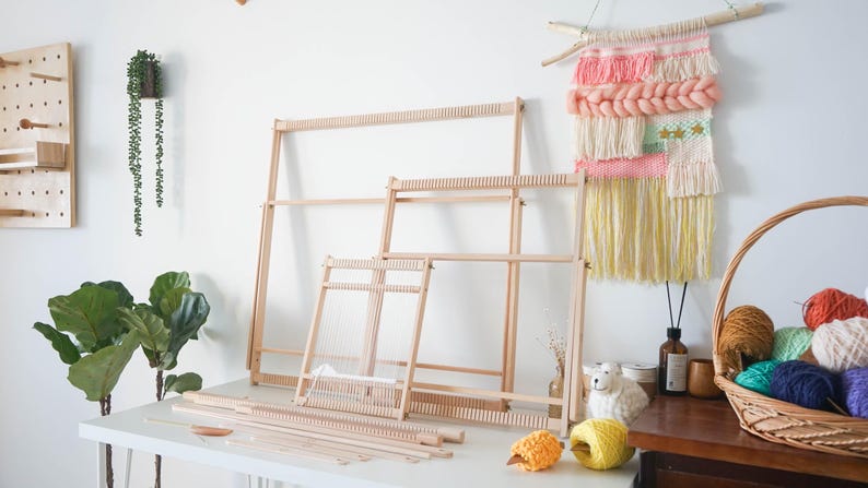 May include: Three light-colored wooden weaving looms of varying sizes are arranged on a white surface. The looms feature vertical notches along the top and bottom bars for threading. Several wooden weaving tools and balls of yarn are placed nearby.