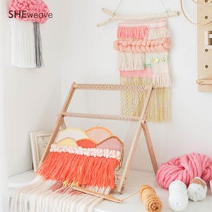 May include: A wooden weaving loom holding a partially completed tapestry with coral, yellow, and white yarn. The tapestry features a mountain-like design with fringe. A finished wall hanging with pink, white, and gold stars hangs in the background.