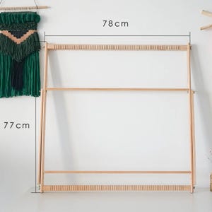 May include: A light-colored wooden weaving loom, 78 cm wide and 77 cm tall, with a rectangular frame and multiple horizontal bars. The loom is designed for textile art and craft projects. A green and beige woven wall hanging is visible on the left.