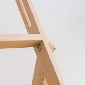 May include: Close-up of a wooden clothing rack with a natural finish. The rack features a simple, minimalist design with angled legs and a horizontal bar. Brass hardware secures the joints. The background is a soft, neutral color.