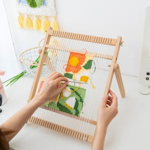 Op de afbeelding: Een houten weefgetouw met een kleurrijk, geometrisch tapijt in wording. Het tapijt heeft oranje, groene en gele vierkanten. Het weefgetouw staat op een wit oppervlak, met een afgewerkte wandhanger op de achtergrond.