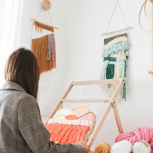 May include: A woman working on a weaving project with a wooden loom. The loom is set up with a colorful woven piece in progress. Two finished wall hangings are displayed on the wall. Various yarn colors are visible, including pink, white, and orange.