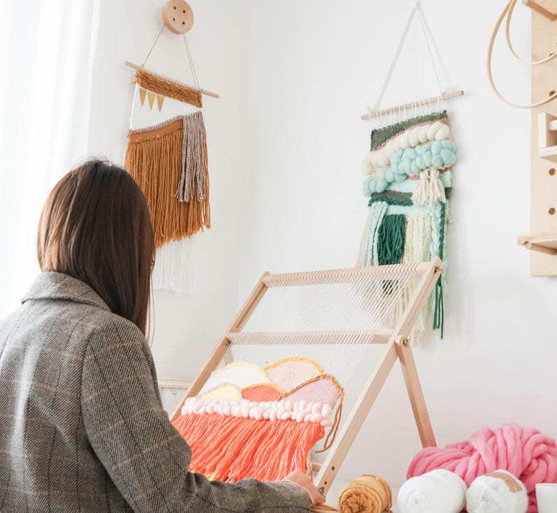 May include: A woman working on a weaving project with a wooden loom. The loom is set up with a colorful woven piece in progress. Two finished wall hangings are displayed on the wall. Various yarn colors are visible, including pink, white, and orange.