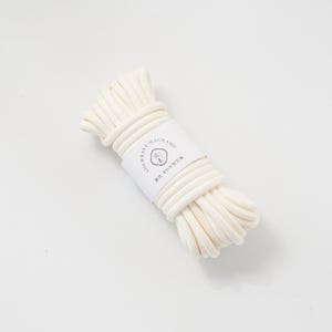 May include: A bundle of off-white macrame cord, tightly wound and secured with a white label. The label features text and a small illustration. The cord appears to be approximately 8mm in diameter, suitable for various craft projects.