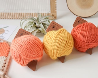 May include: Three skeins of yarn in shades of orange and yellow are displayed. Each yarn ball has a brown tag attached. The word "SHEweave" is visible in the upper left corner. A small wooden loom and a plant are in the background.