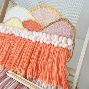 May include: A woven wall hanging on a wooden loom featuring a mountain landscape design in yellow, white, orange, and pink yarn. The piece includes a row of thick, pale pink braided yarn and a long fringe of bright orange and white yarn strands.