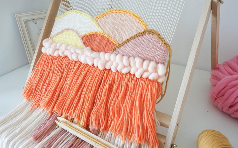 May include: A woven wall hanging on a wooden loom featuring a mountain landscape design in yellow, white, orange, and pink yarn. The piece includes a row of thick, pale pink braided yarn and a long fringe of bright orange and white yarn strands.