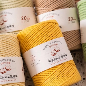 Puede incluir: Primer plano de varios carretes de cordón de algodón de 2 mm en varios colores, incluyendo amarillo, beige y marrón. Cada carrete tiene una etiqueta con texto en inglés y chino. Ideal para manualidades.