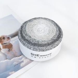 May include: A round skein of yarn with a gradient color scheme, transitioning from dark gray to silver and white. The yarn is labeled "SHEweave" with Chinese characters. The yarn is sitting on a white surface next to a magazine.