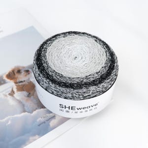 May include: A skein of yarn with a gradient of black, silver, and white. The yarn is wound into a round shape and is on a white label with the text "SHEweave". The image also includes a picture of a dog.