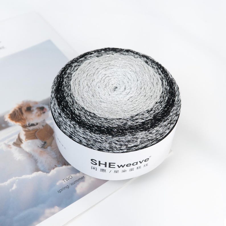 May include: A skein of yarn with a gradient of black, silver, and white. The yarn is wound into a round shape and is on a white label with the text "SHEweave". The image also includes a picture of a dog.