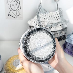 May include: A close-up of a skein of yarn with a gradient of black, white, and gray. The yarn is held in front of a crocheted handbag with a pearl handle. Other skeins of yarn in yellow and blue are also visible.