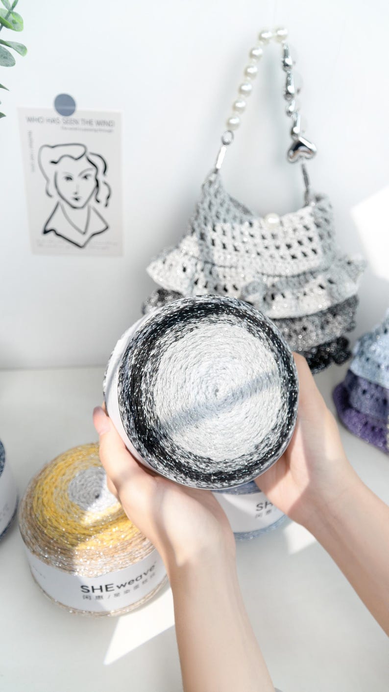 May include: A close-up of a skein of yarn with a gradient of black, white, and gray. The yarn is held in front of a crocheted handbag with a pearl handle. Other skeins of yarn in yellow and blue are also visible.