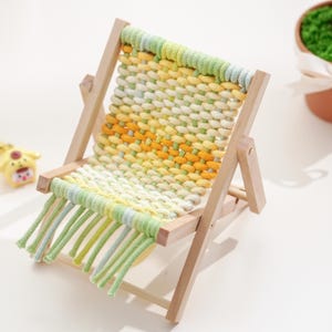 May include: A miniature wooden folding chair with a woven seat. The seat is made of yellow, green, and white yarn. The chair has a light wood frame and green fringe.