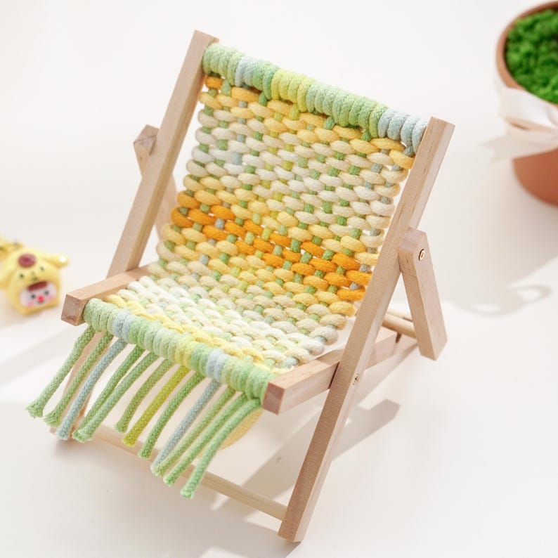 May include: A miniature wooden folding chair with a woven seat. The seat is made of yellow, green, and white yarn. The chair has a light wood frame and green fringe.