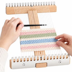 Op de afbeelding: Een houten weefgetouw met metalen tanden aan de boven- en onderkant, voorzien van witte verticale scheringdraden en een gedeeltelijk geweven textiel met horizontale strepen in lichtroze, saliegroen en lichtblauw, bewerkt met een zwarte naald.