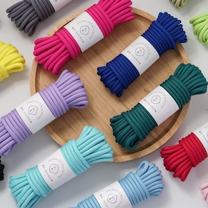 Puede incluir: Una colección de paquetes de cuerda de macramé multicolor, cada uno envuelto con una banda de papel blanco con un logotipo circular y texto. Las cuerdas se presentan en varios tonos vibrantes como rosa, azul, amarillo, verde, morado, negro y blanco.