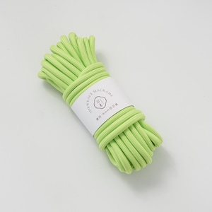 May include: A bundle of light green macrame cord, wrapped with a white label. The label features the text "SHEWEAVE-MACRAME" and other text in a different language. The cord is tightly wound, showcasing its texture and color.