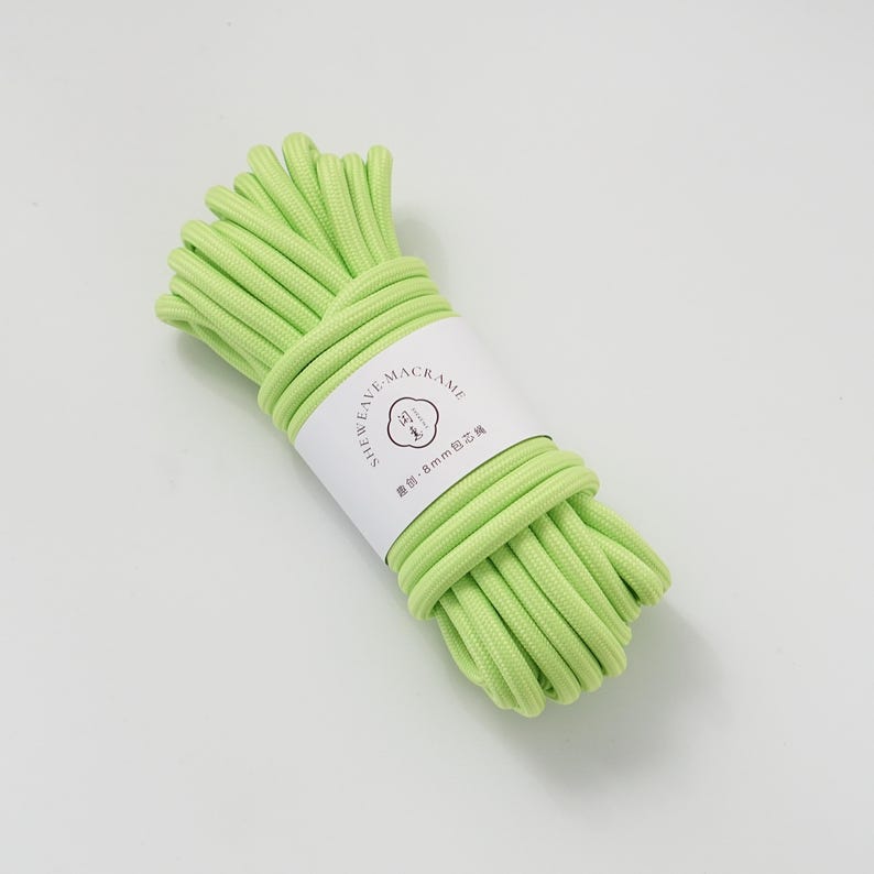 May include: A bundle of light green macrame cord, wrapped with a white label. The label features the text "SHEWEAVE-MACRAME" and other text in a different language. The cord is tightly wound, showcasing its texture and color.