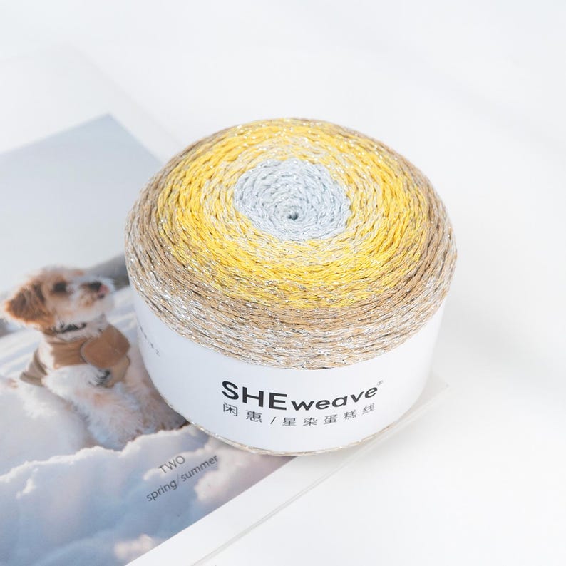 May include: A ball of yarn with a gradient of colors, including yellow, tan, and silver. The yarn is wrapped around a white cylinder with the brand name "SHEweave" printed on it. The image also includes the text "TWO spring/summer".