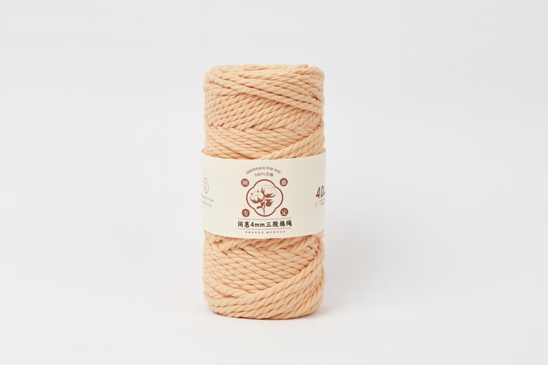 May include: A cylindrical spool of peach-colored cotton cord wrapped in a paper band. The band features a central circular logo with a cotton plant illustration, the text "4mm", and Chinese characters. The cord has a thick, twisted texture.