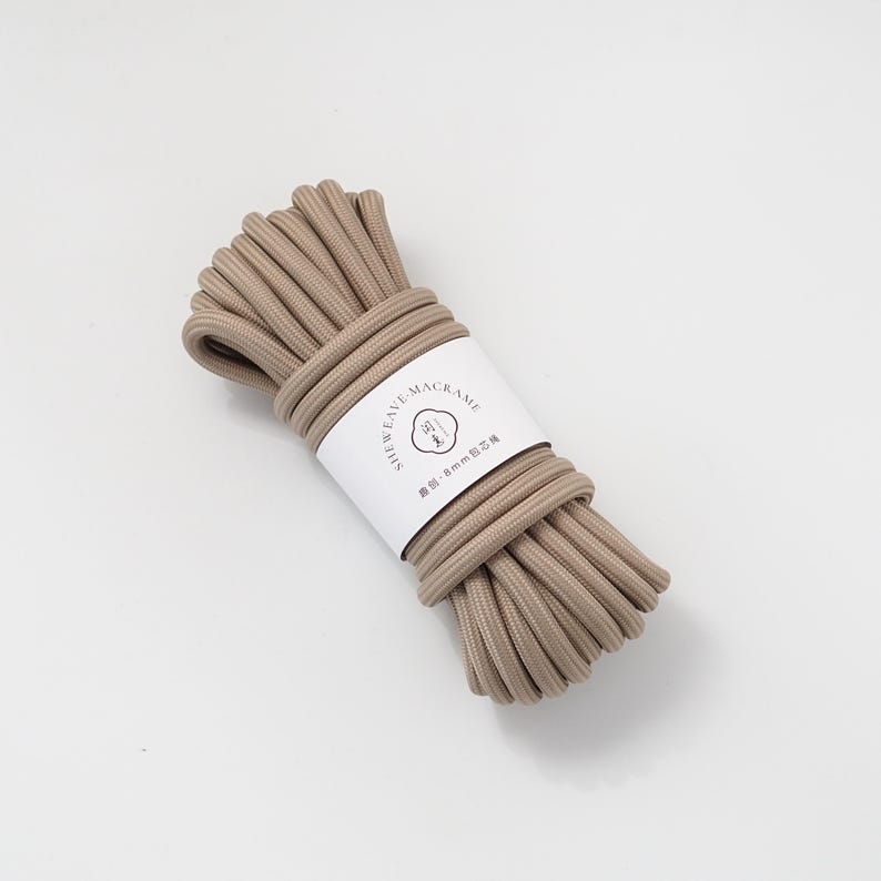 May include: A bundle of beige macrame cord, tightly wound and secured with a white label. The label features the text "SHEAVE MACRAME" and additional text in a different language. The cord appears to be approximately 8mm in diameter.